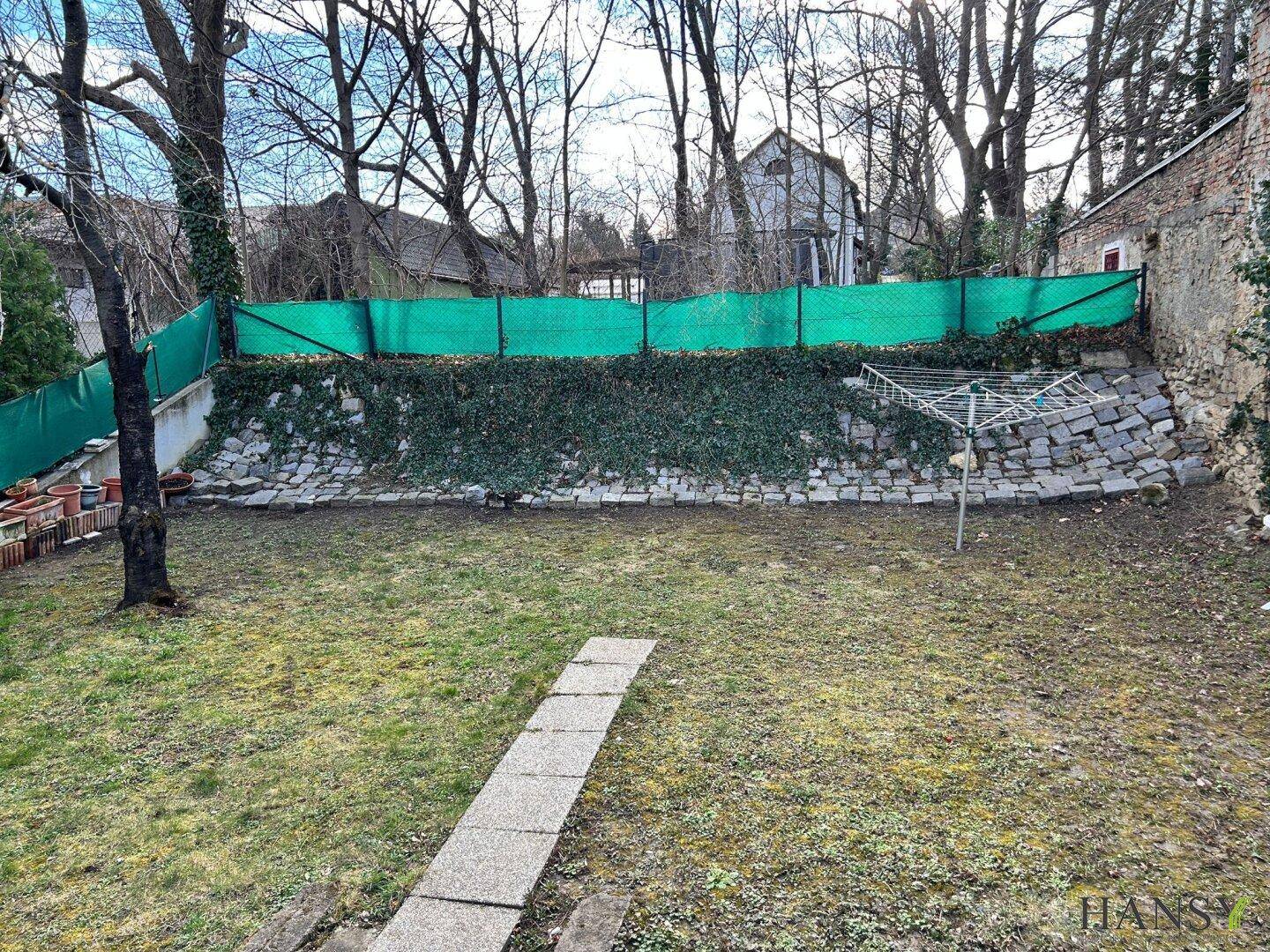 Garten mit Rasenfläche, bewachsenem Hang und Wäscheleine, umgeben von Bäumen und einer Mauer.