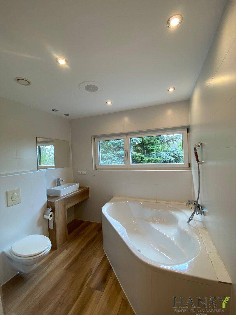 Helles Badezimmer mit Badewanne, Einzelwaschbecken, Toilette und Fenster mit Blick ins Grüne.