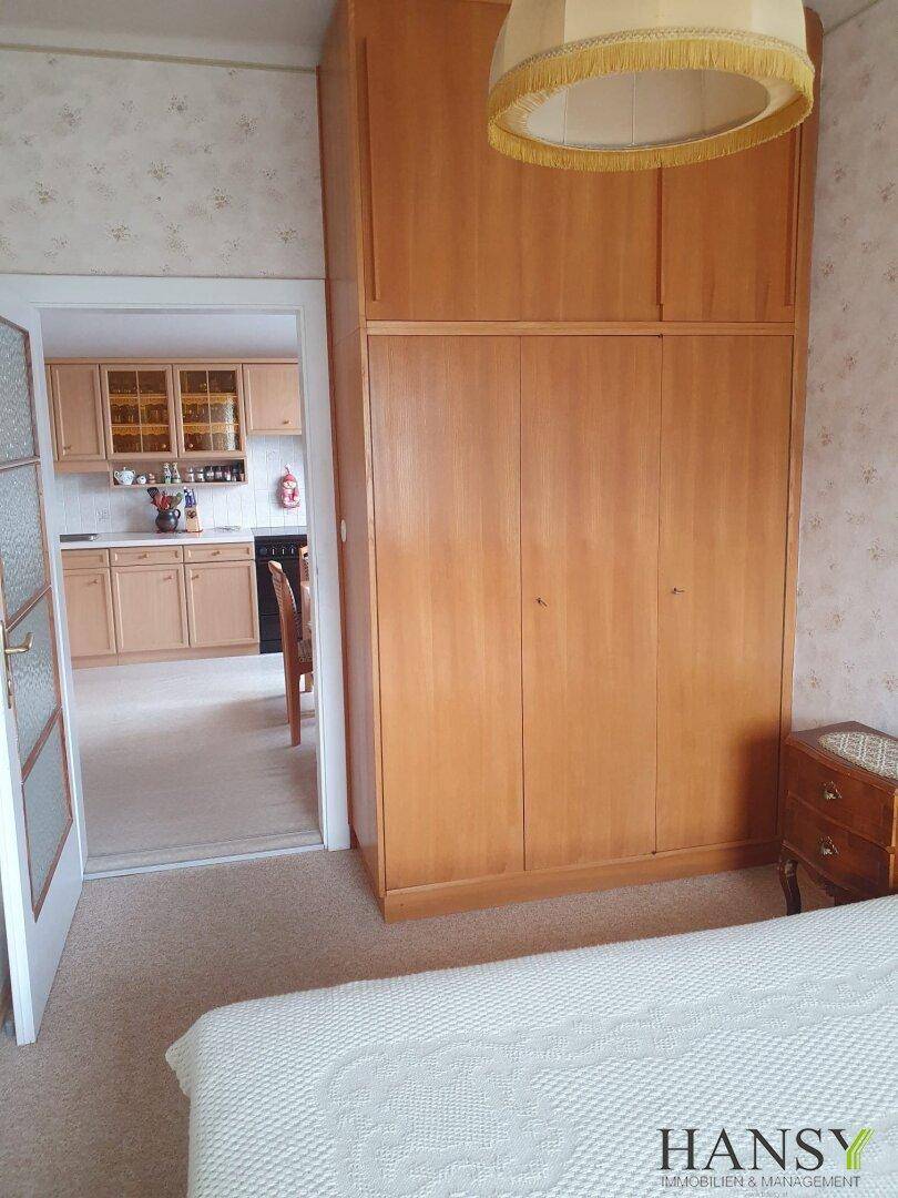 Schlafzimmer mit großem Holzschrank und gemusterter Tapete, Blick in die angrenzende Küche.