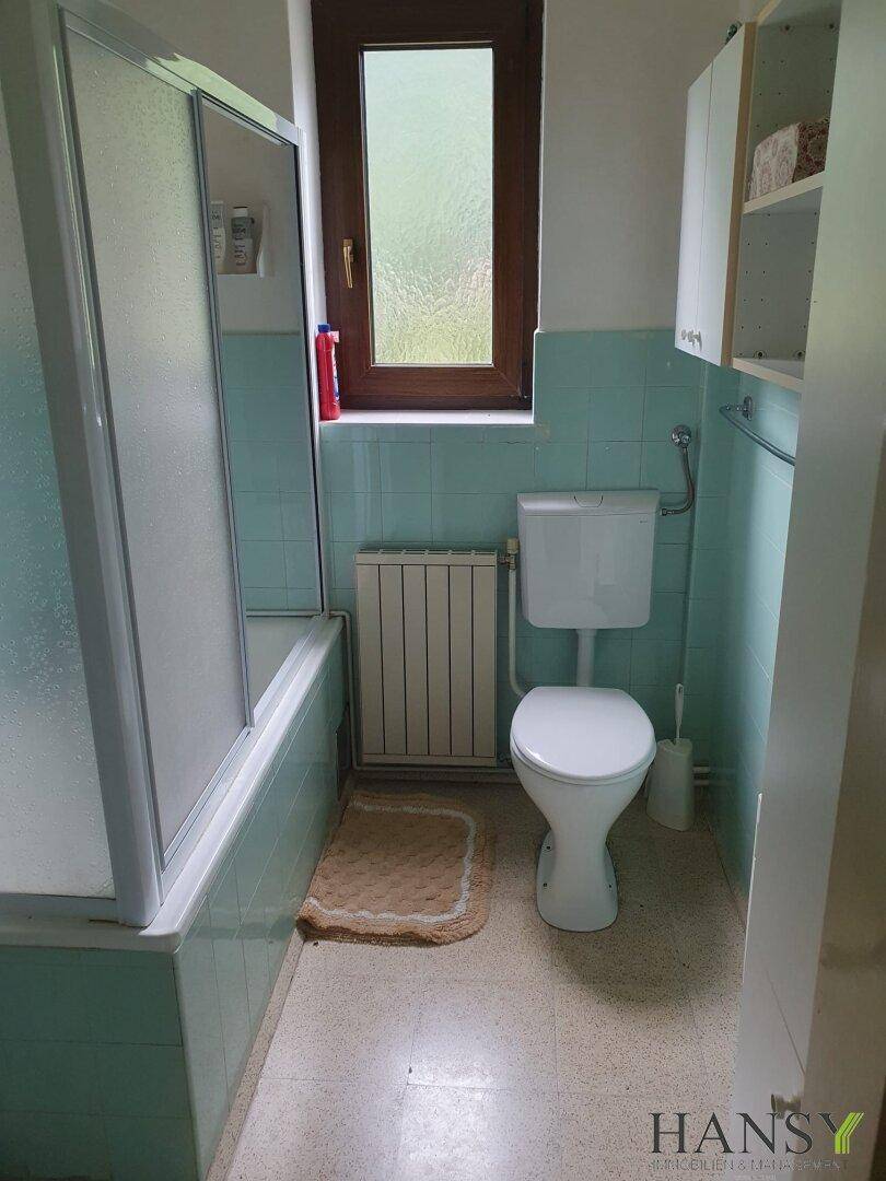 Badezimmer mit hellblauen Fliesen, Badewanne mit Duschwand, Toilette und einem Fenster für Tageslicht.