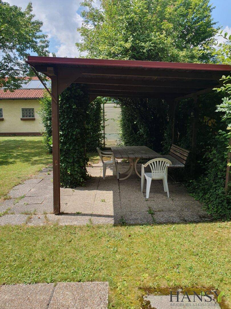 Gemütlicher Sitzbereich im Garten unter einer Holzpergola, umgeben von üppigem Grün.