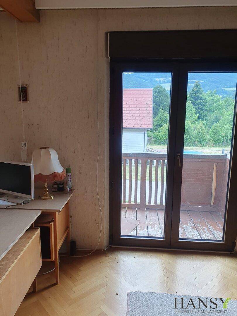 Zimmer mit Schreibtisch und gemusterter Tapete, Zugang zu einem Balkon mit weitem Blick ins Grüne.