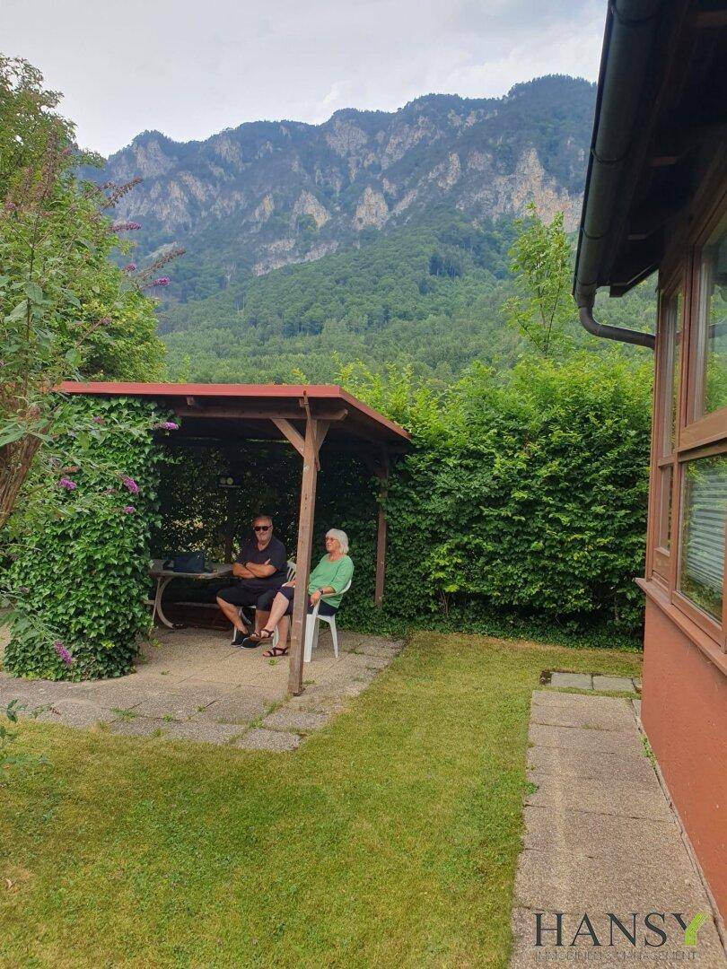 Personen entspannen im Garten unter einer Pergola, mit herrlichem Blick auf die umliegenden Berge.