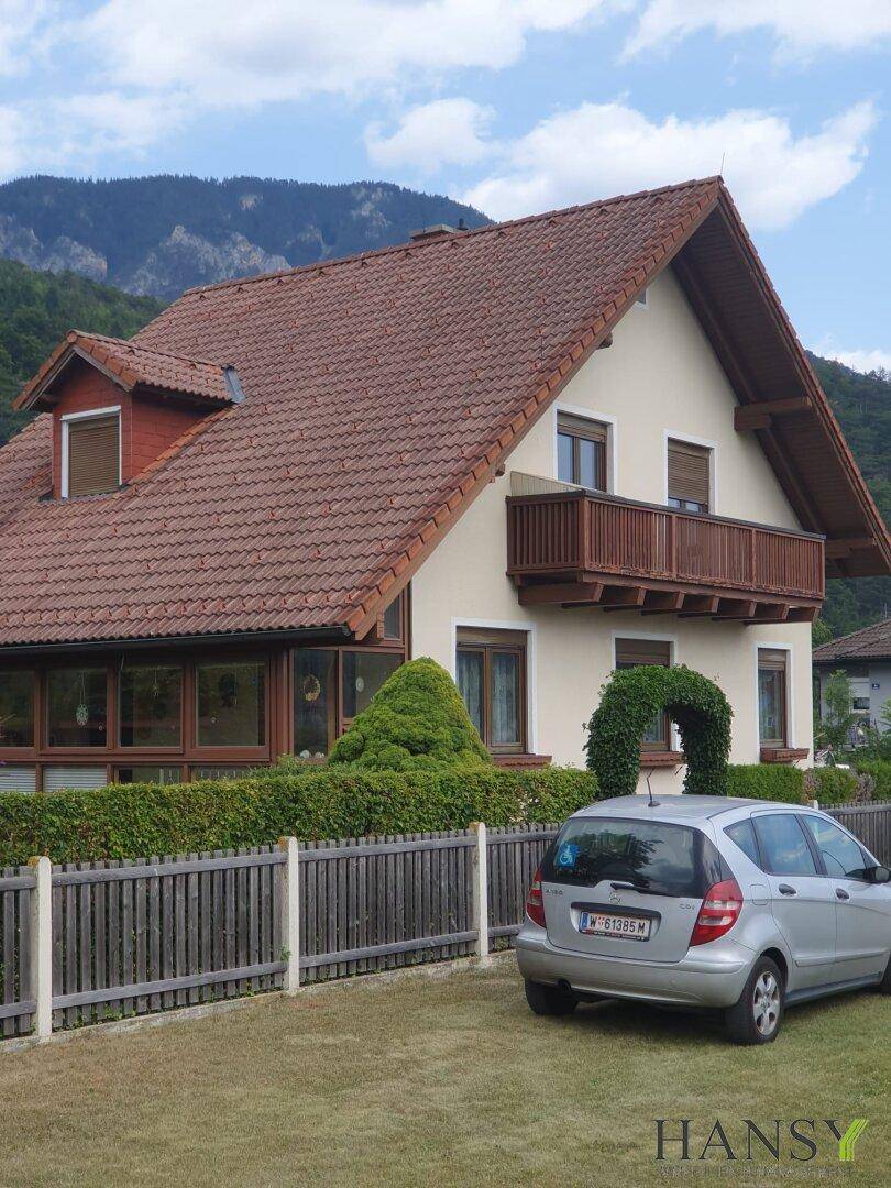 Seitenansicht des Hauses mit rotem Ziegeldach, Gaube und einem gepflegten Garten vor bergiger Kulisse.
