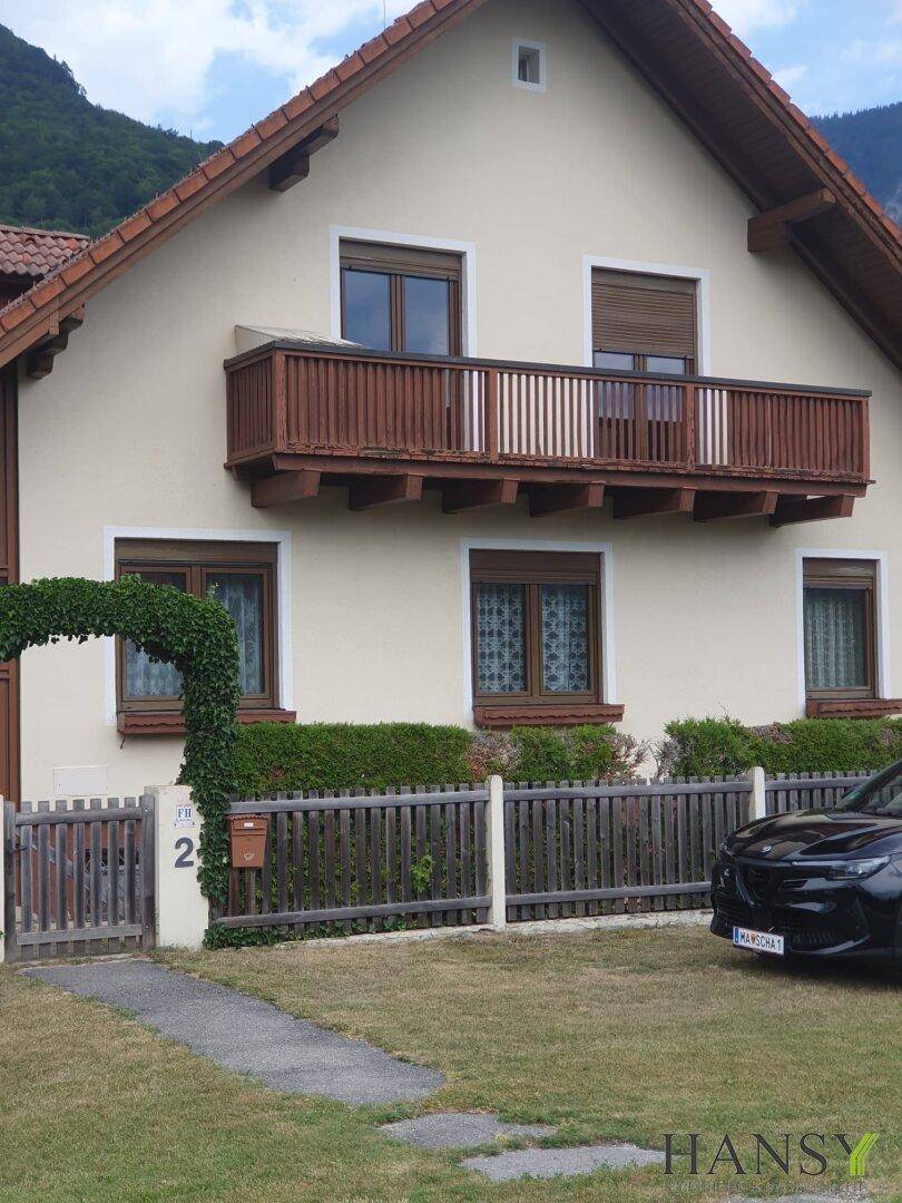 Detailansicht der Hausfassade mit Holzbalkon, Fenstern mit Rollläden und einem begrünten Eingangsbereich.
