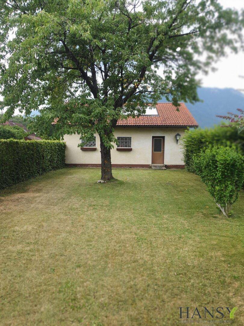 Kleines Nebengebäude oder Gartenhaus auf einer Rasenfläche, umgeben von einer dichten Hecke.