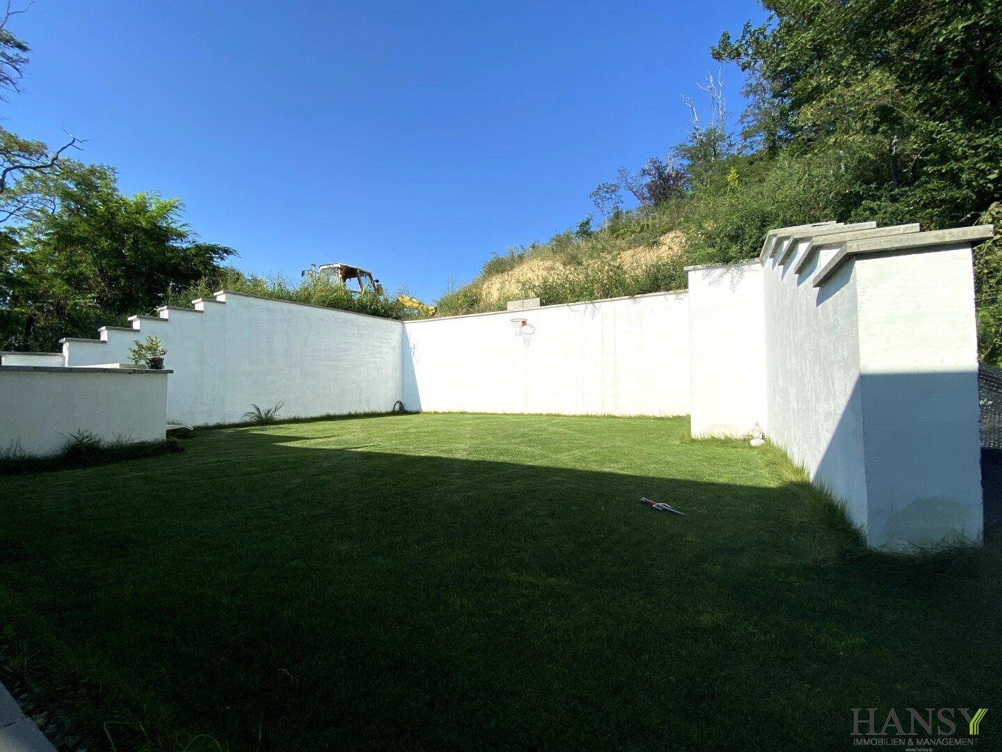 Gepflegter Gartenbereich mit Rasenfläche, umgeben von weißen Mauern und natürlicher Vegetation.