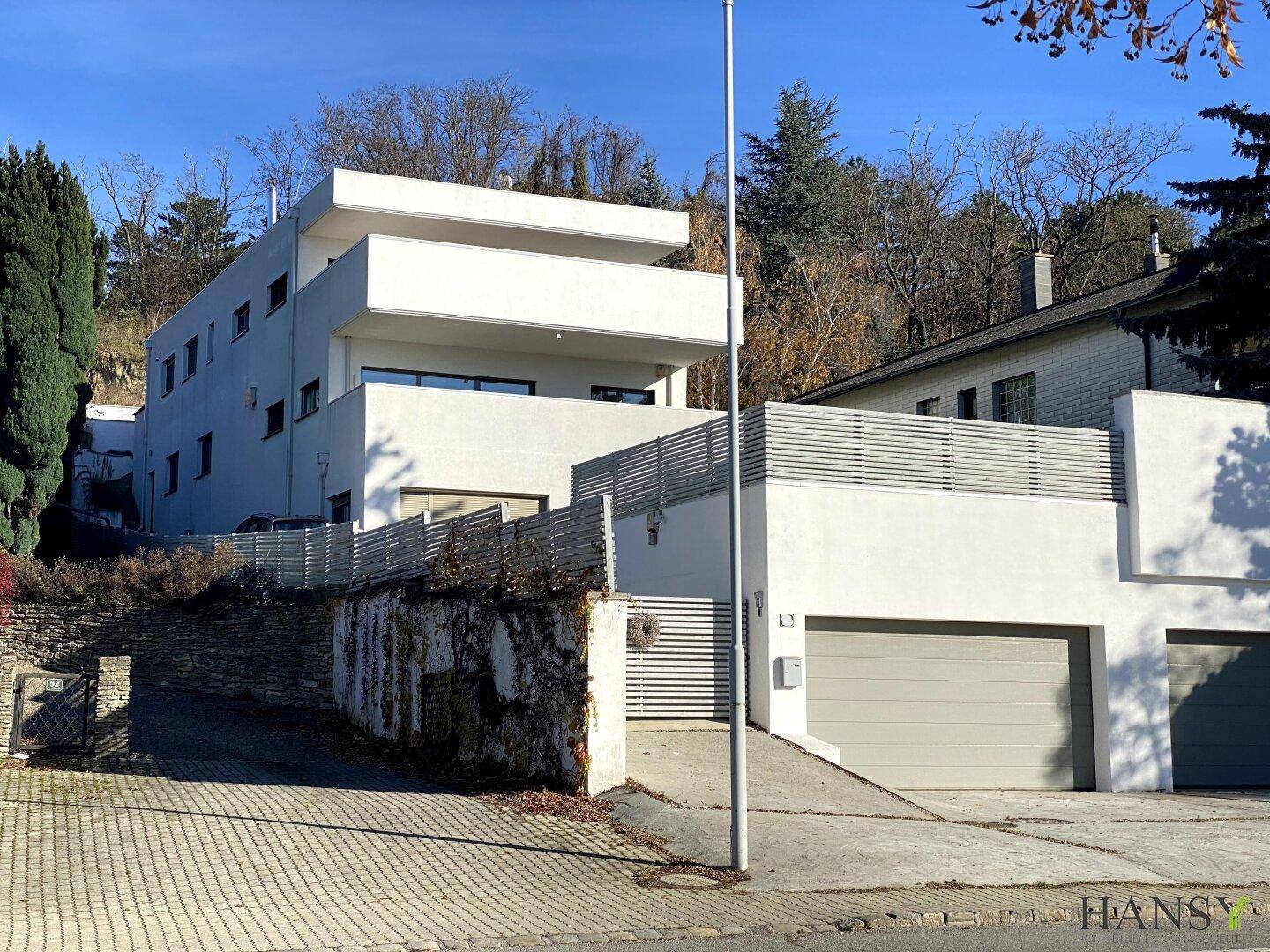 Zeitgenössisches weißes Gebäude mit klaren Linien und Garagenzufahrt, eingebettet in eine grüne Hügellandschaft.