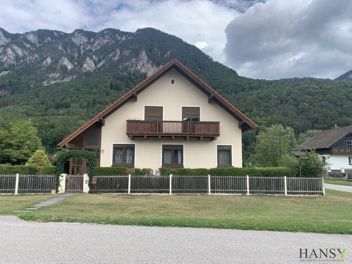 Charmantes Haus mit Balkon und gepflegtem Vorgarten vor majestätischen Bergen und viel Grün.