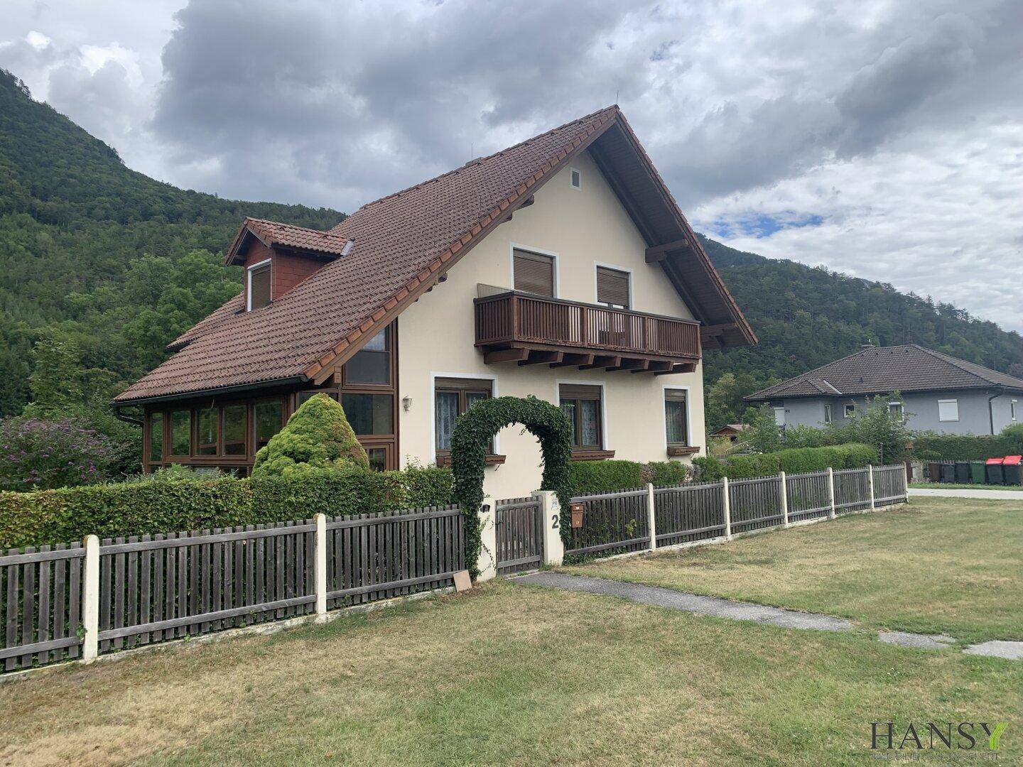 Freistehendes Haus mit Balkon und Wintergarten, umgeben von einem grünen Garten und Bergen.