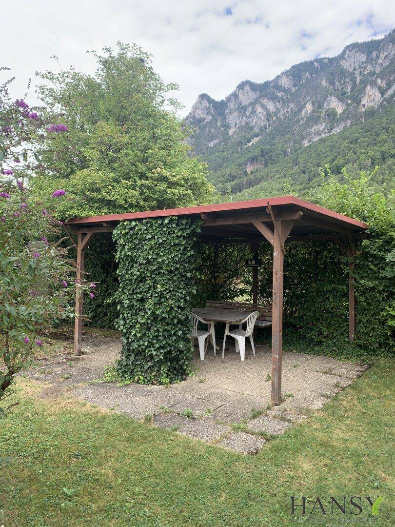 Überdachter Sitzbereich im Garten mit Tisch und Stühlen, umgeben von viel Grün und Bergen.