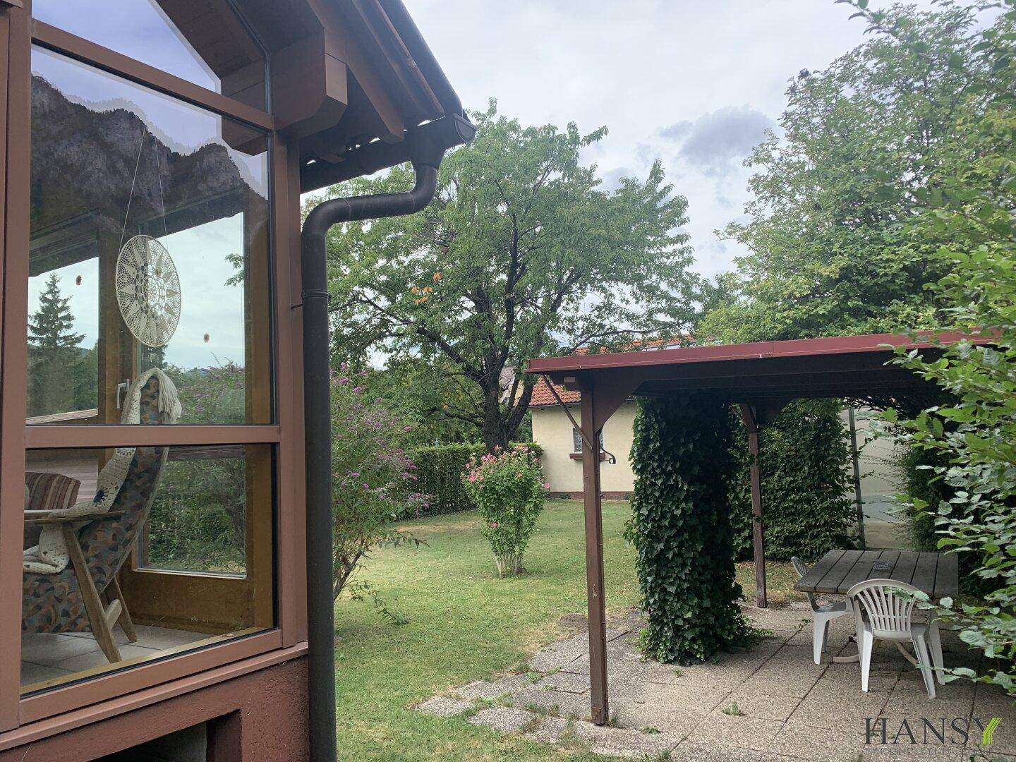 Grüner Gartenbereich mit überdachtem Sitzplatz, Rasenfläche und angrenzendem Wintergarten.
