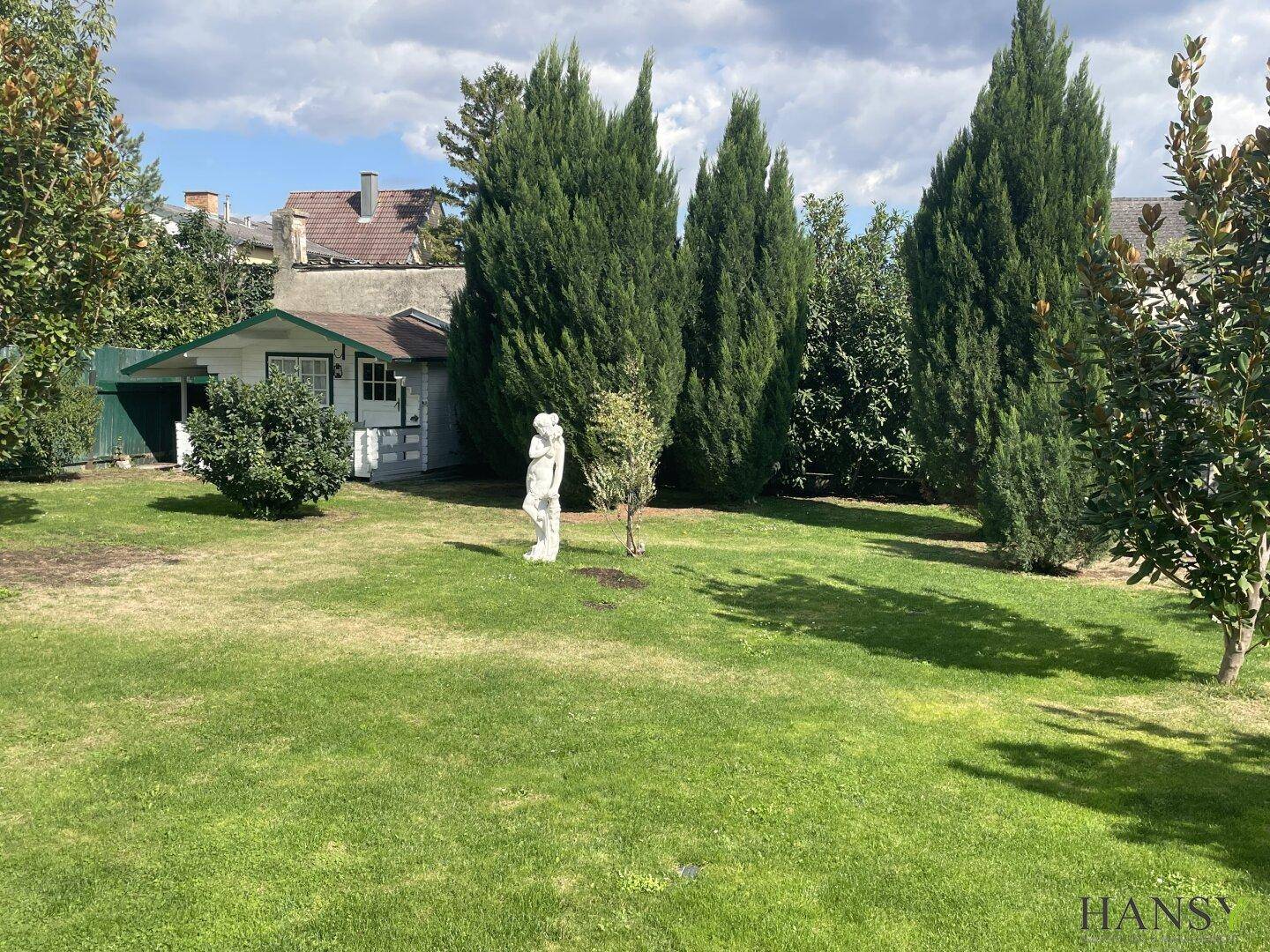 Gepflegter Gartenbereich mit hohen Nadelbäumen, Rasenfläche und einem kleinen Gartenhaus.