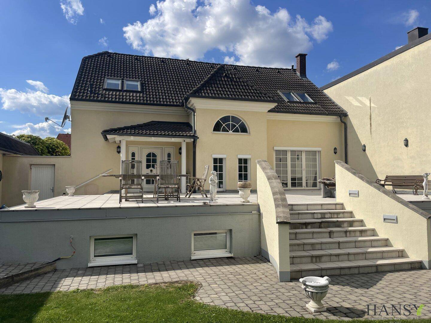 Rückansicht des Hauses mit großer Terrasse, Außentreppe und Gartenbereich an einem sonnigen Tag.
