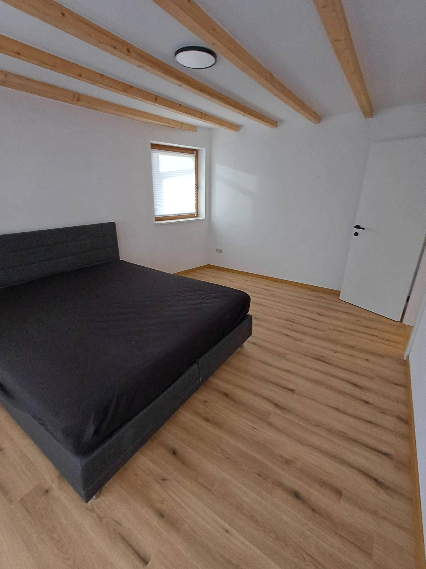 Helles Schlafzimmer mit Holzbalken, Laminatboden und einem Fenster mit Plissee.