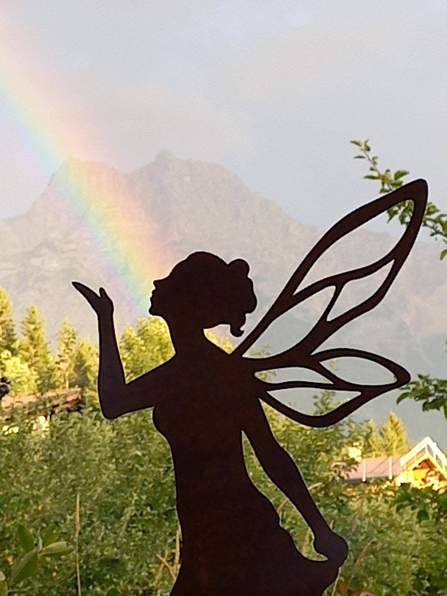 Dekorative Feenfigur im Garten mit einem Regenbogen und Bergen im Hintergrund.