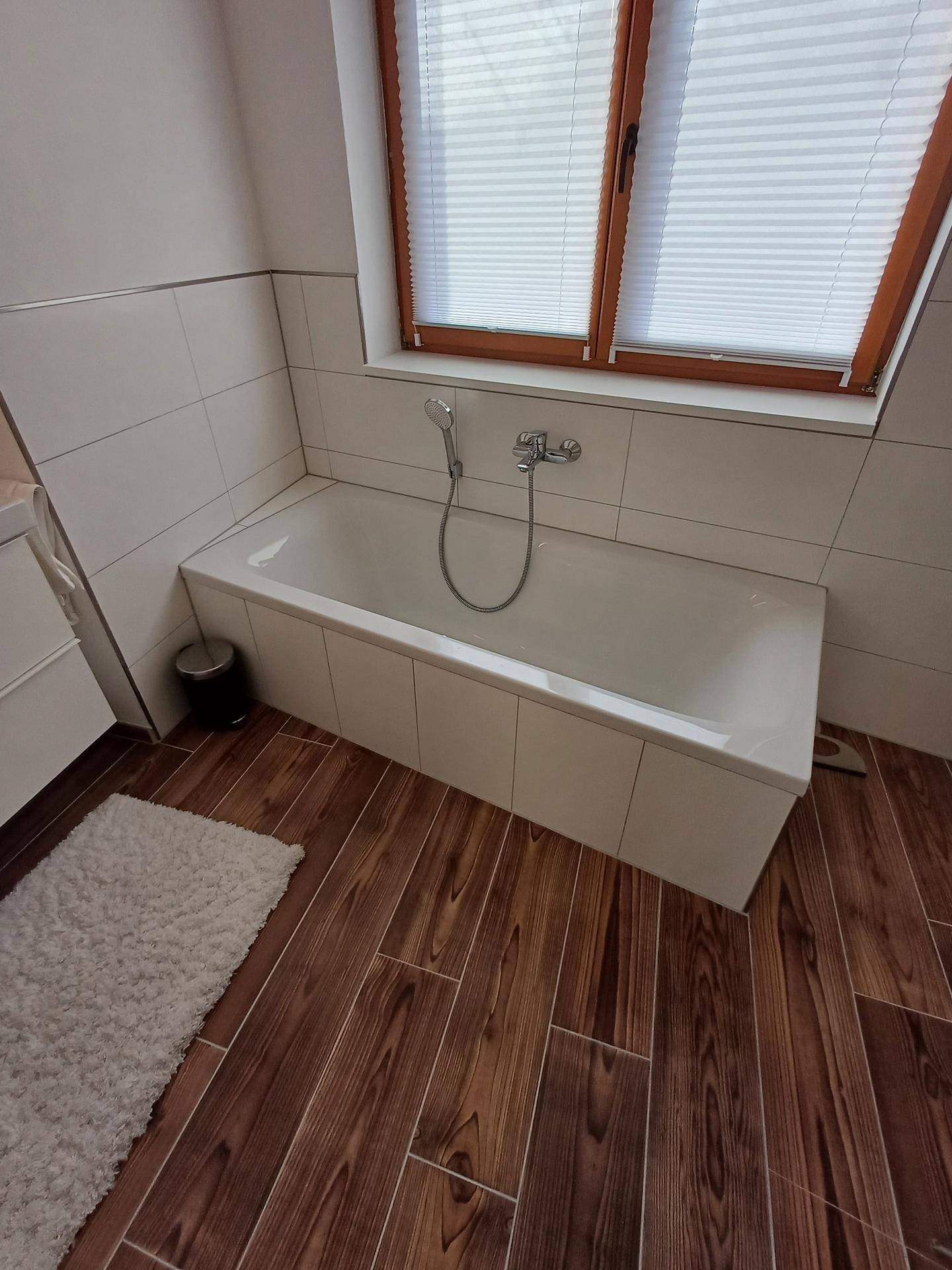 Badewanne unter einem Fenster mit Plissee, umgeben von weißen Fliesen und Holzoptikboden.