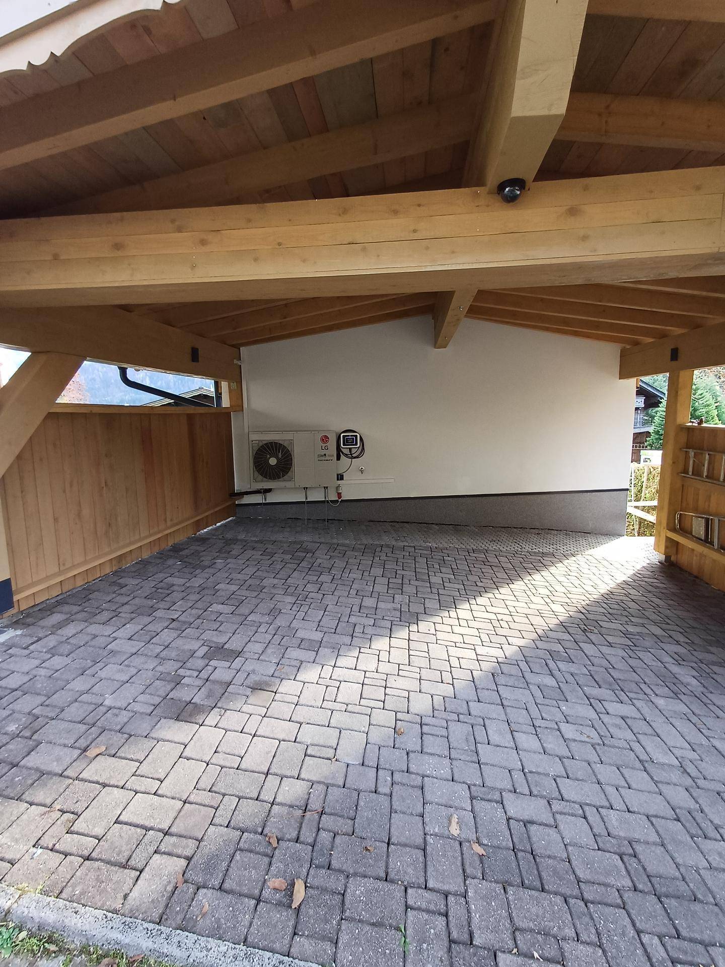 Überdachter Stellplatz mit Pflastersteinen und sichtbarer Wärmepumpe an der Wand.