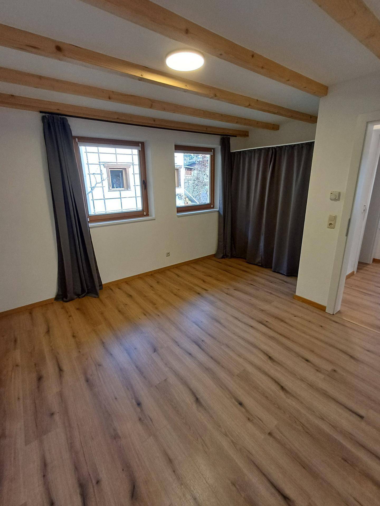 Heller Raum mit Laminatboden, sichtbaren Holzbalken und dunklen Vorhängen.