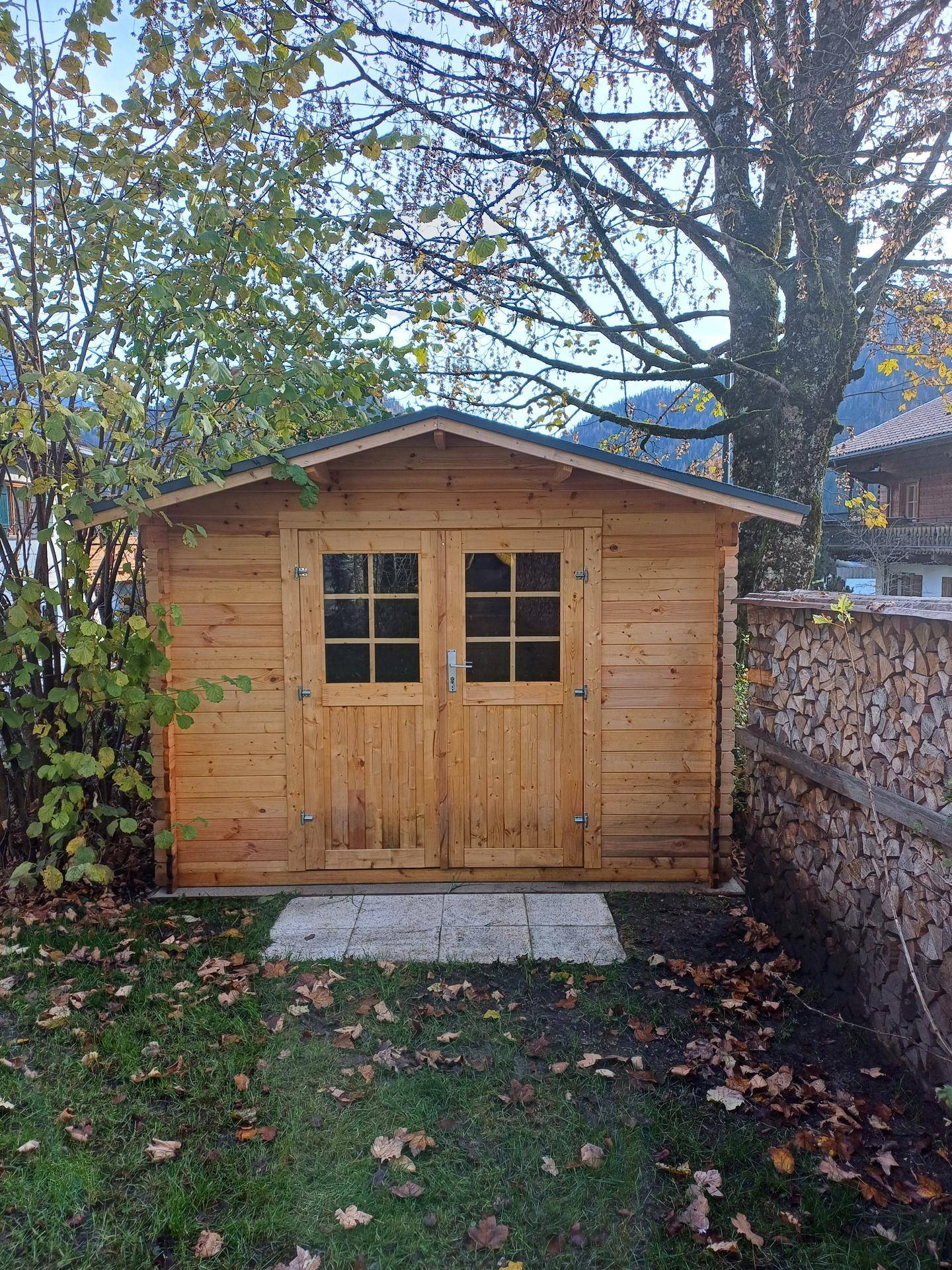 Kleiner Holzschuppen mit Doppeltür und Fenstern im Garten, umgeben von Bäumen.