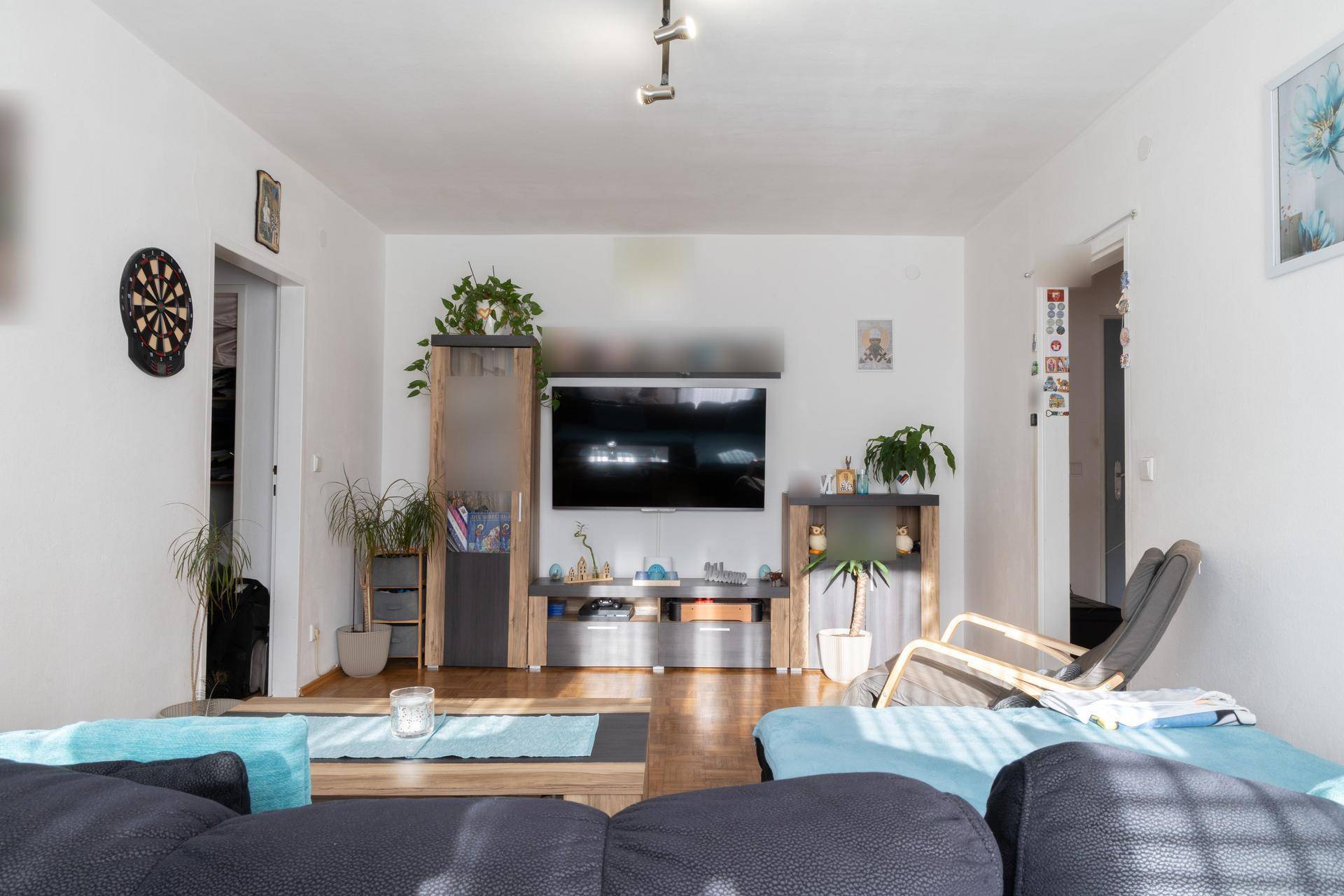 Helles Wohnzimmer mit Fernseher, Sofa und Parkettboden, dekoriert mit Pflanzen und Bildern.