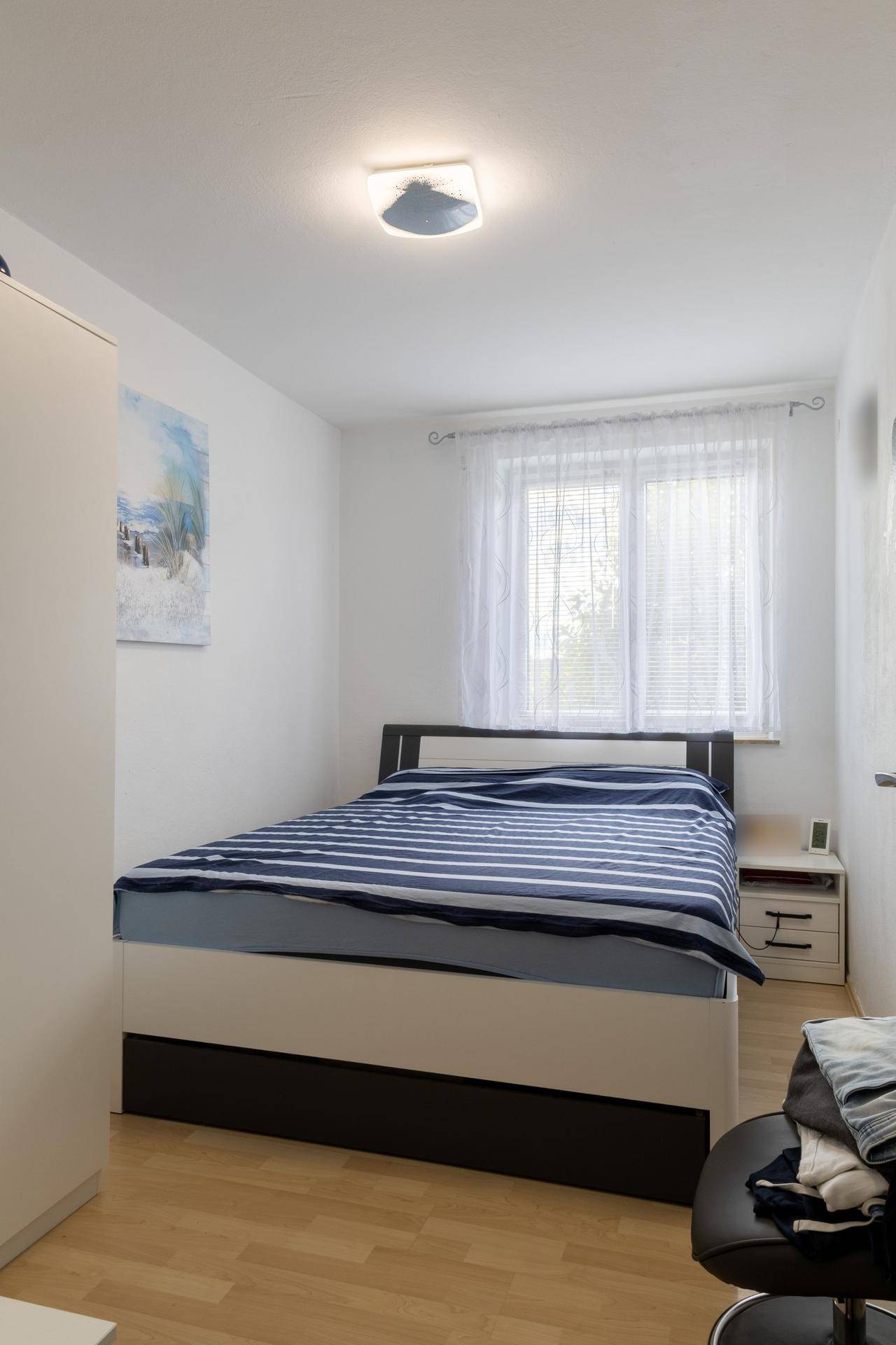 Schlafzimmer mit Doppelbett, weißem Kleiderschrank und Fenster mit Jalousien.