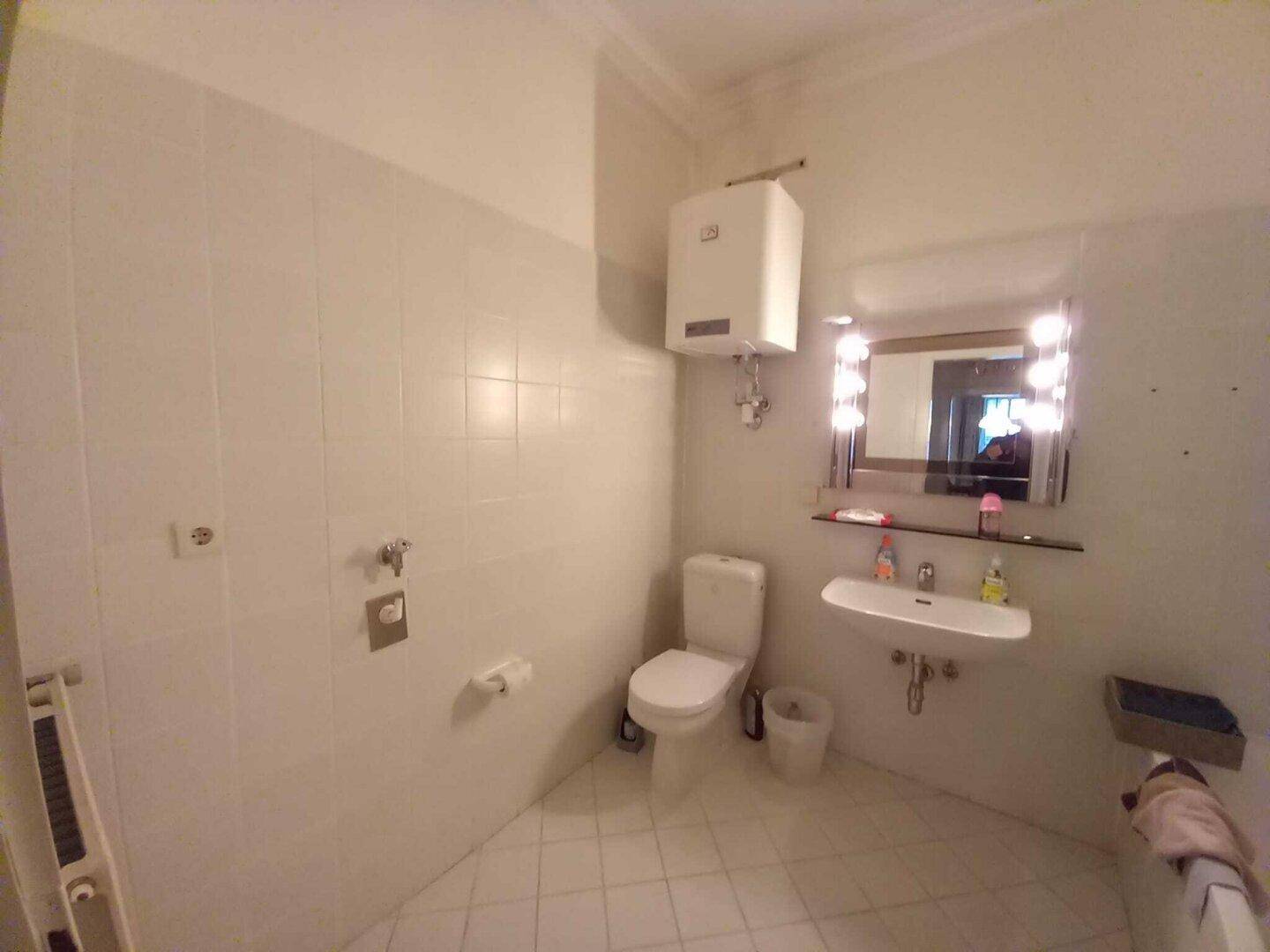 Helles Badezimmer mit weißen Fliesen, Toilette, Waschbecken und beleuchtetem Spiegel.