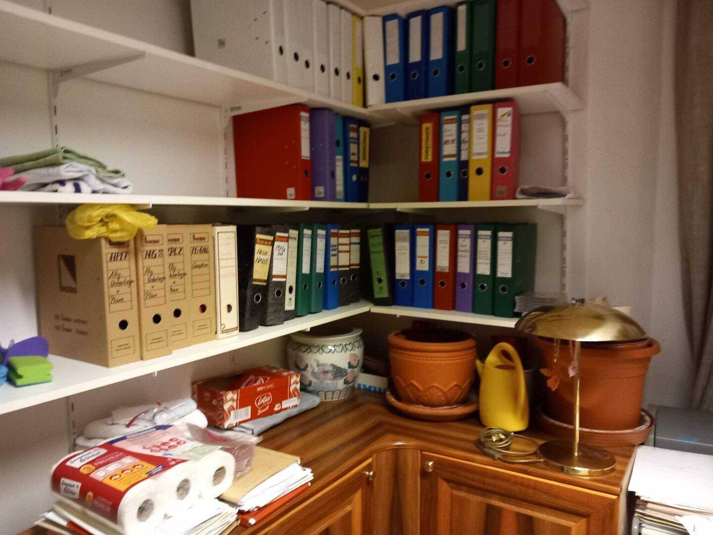 Eckbereich eines Abstellraums mit Regalen, Aktenordnern und Dekorationsgegenständen.