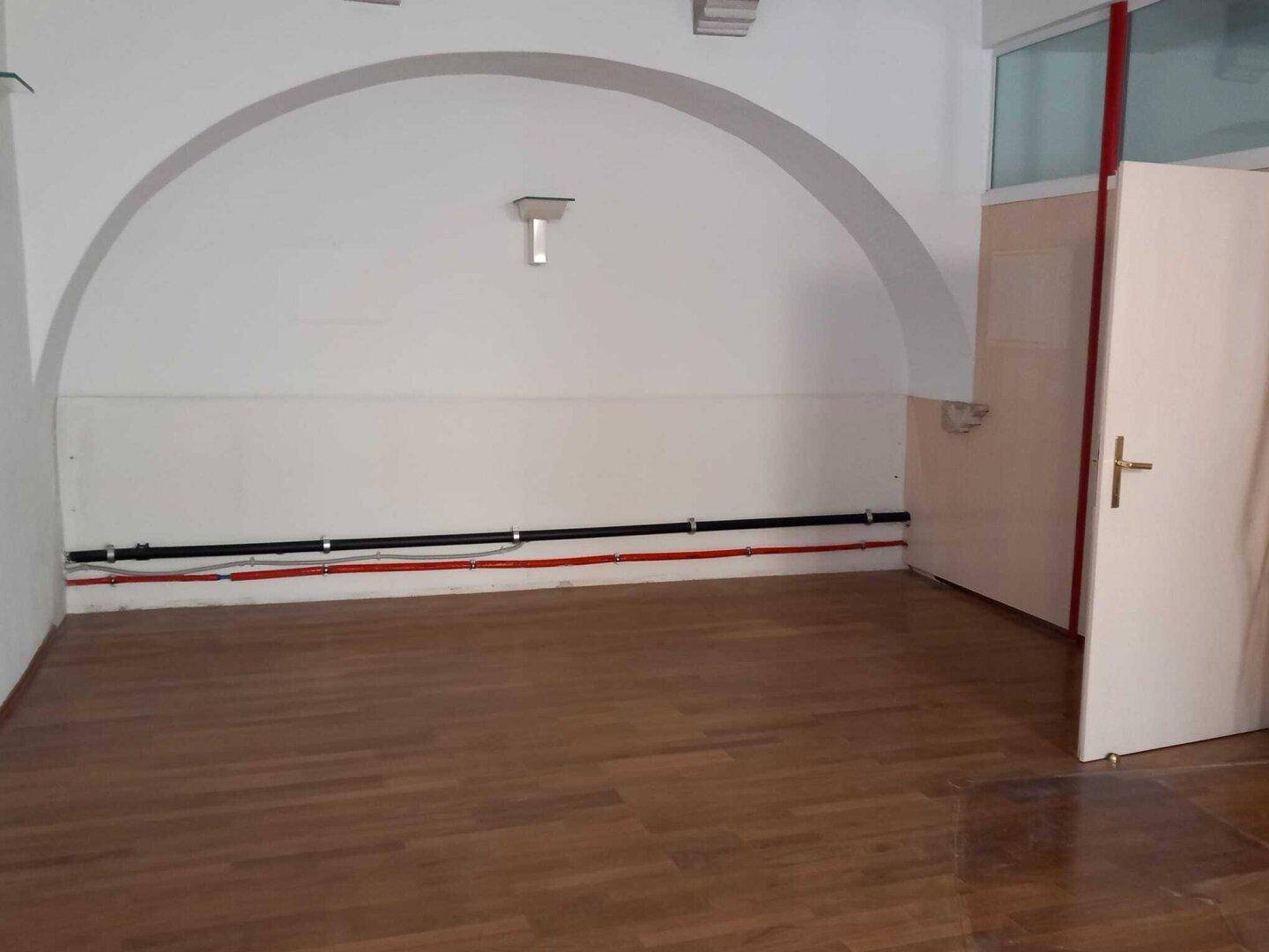 Ein leerer Raum mit Holzboden, einer gewölbten Decke und sichtbaren Leitungen an der Wand.