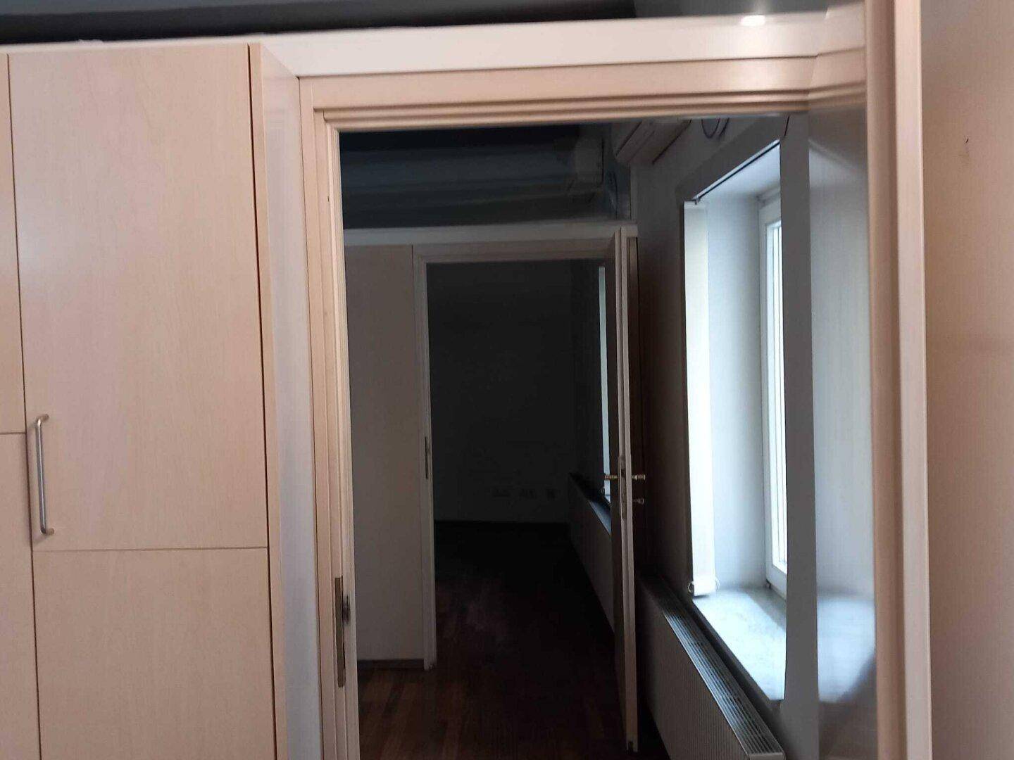 Blick durch eine Tür in einen Flur mit einem Fenster und einem Heizkörper, umgeben von hellen Schränken.