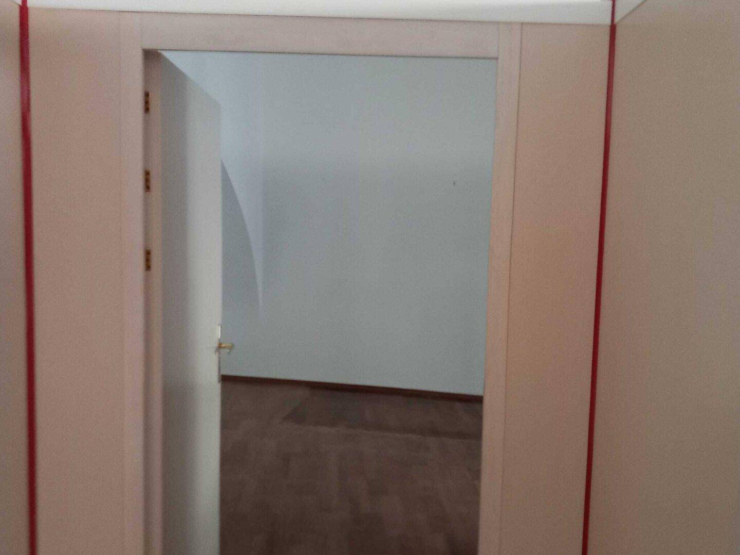 Blick durch eine offene Tür in einen leeren Raum mit hellem Boden und weißen Wänden.