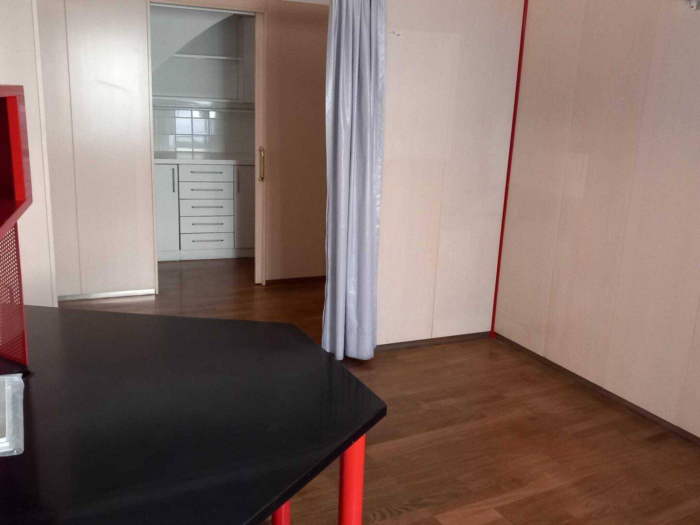 Ein Raum mit Holzboden und einem schwarzen Schreibtisch, der zu einer Schiebetür mit Blick auf eine Küchenzeile führt.