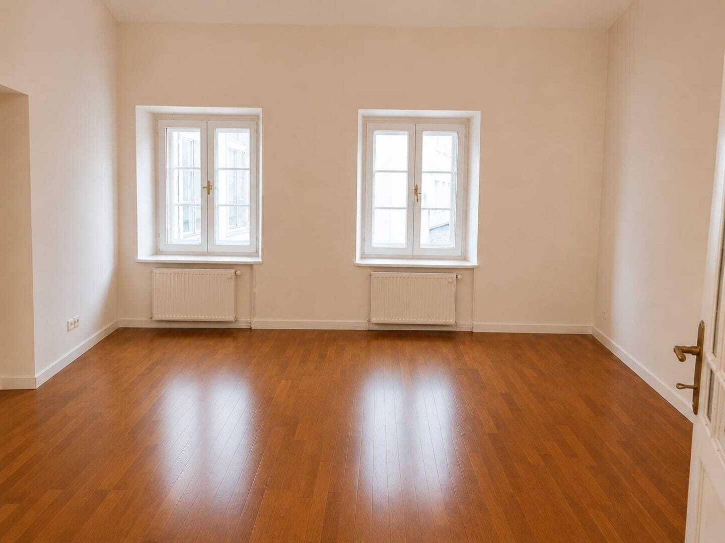 Geräumiges Zimmer mit dunklem Holzfußboden, hellen Wänden und zwei klassischen Fenstern mit Heizkörpern.