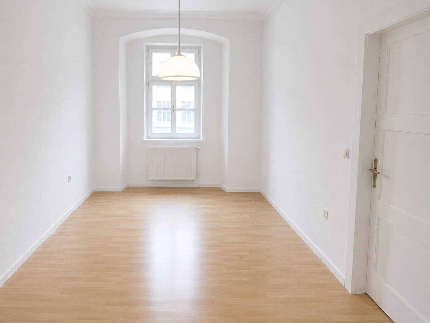 Helles Zimmer mit hellem Holzfußboden, weißen Wänden und einem Fenster mit Heizkörper darunter.