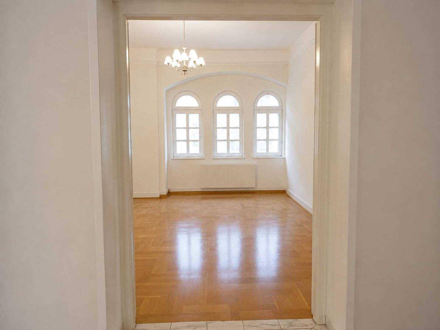 Blick durch einen Türrahmen in ein helles Zimmer mit Fischgrätparkett und eleganten Rundbogenfenstern.