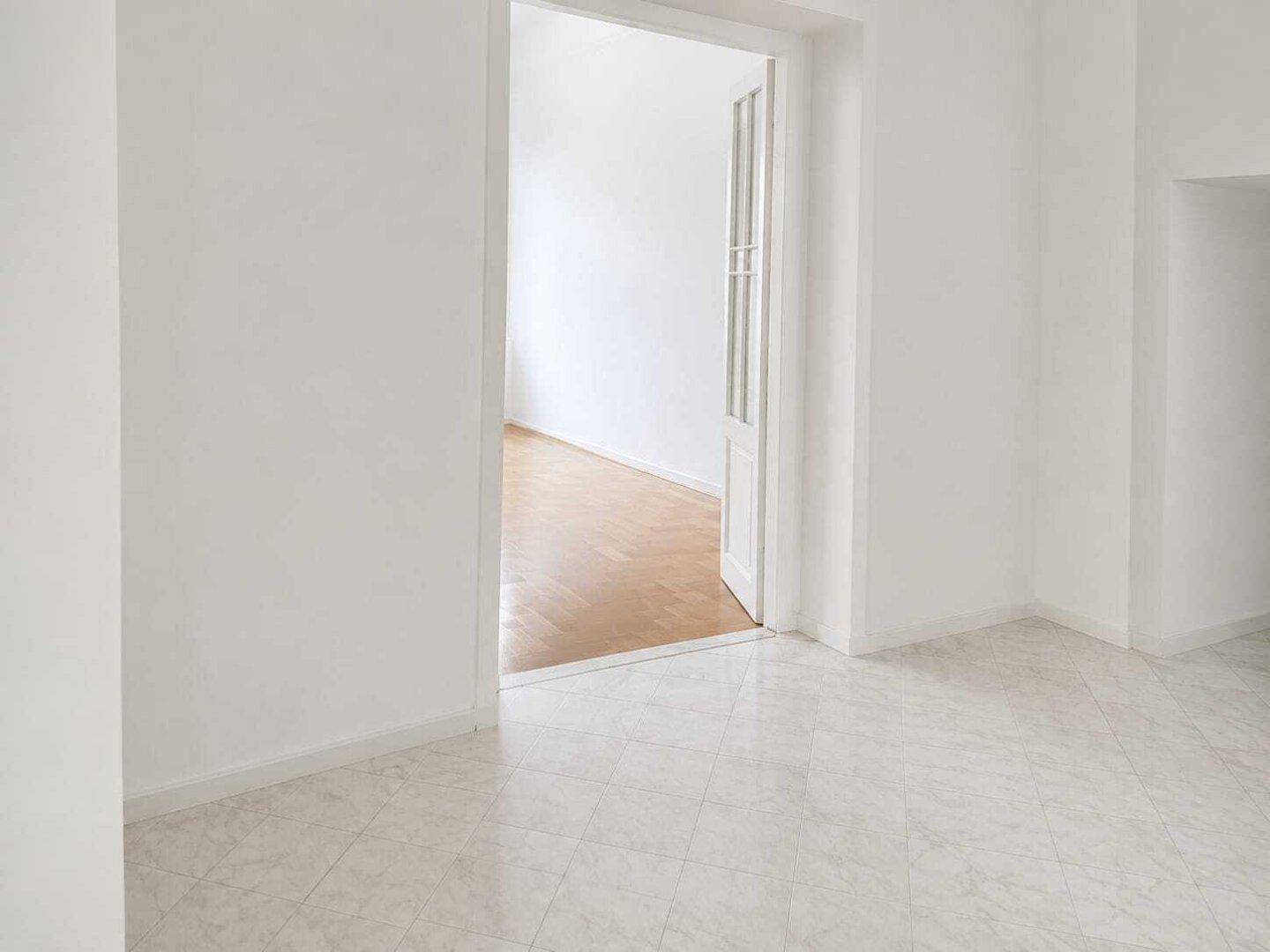 Flur mit hellem Fliesenboden und Blick durch eine Glastür in einen angrenzenden Raum mit Holzfußboden.