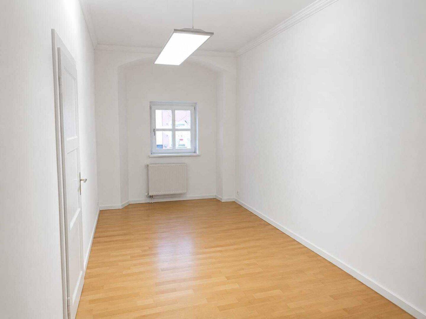 Schlichtes Zimmer mit hellem Holzfußboden, weißen Wänden und einem Fenster, ausgestattet mit moderner Deckenleuchte.
