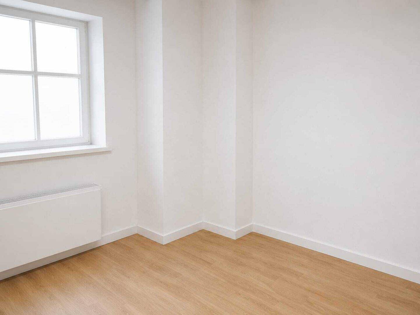 Ecke eines hellen Zimmers mit hellem Holzfußboden, weißen Wänden und einem Fenster mit Heizkörper.