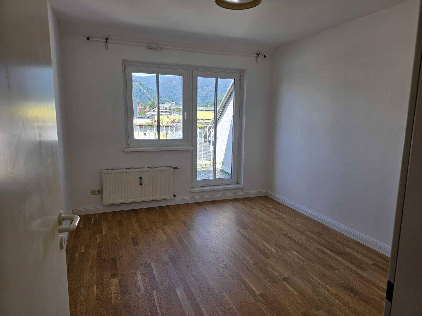 Helles Zimmer mit Holzboden, Fenster und Zugang zu einem kleinen französischen Balkon.