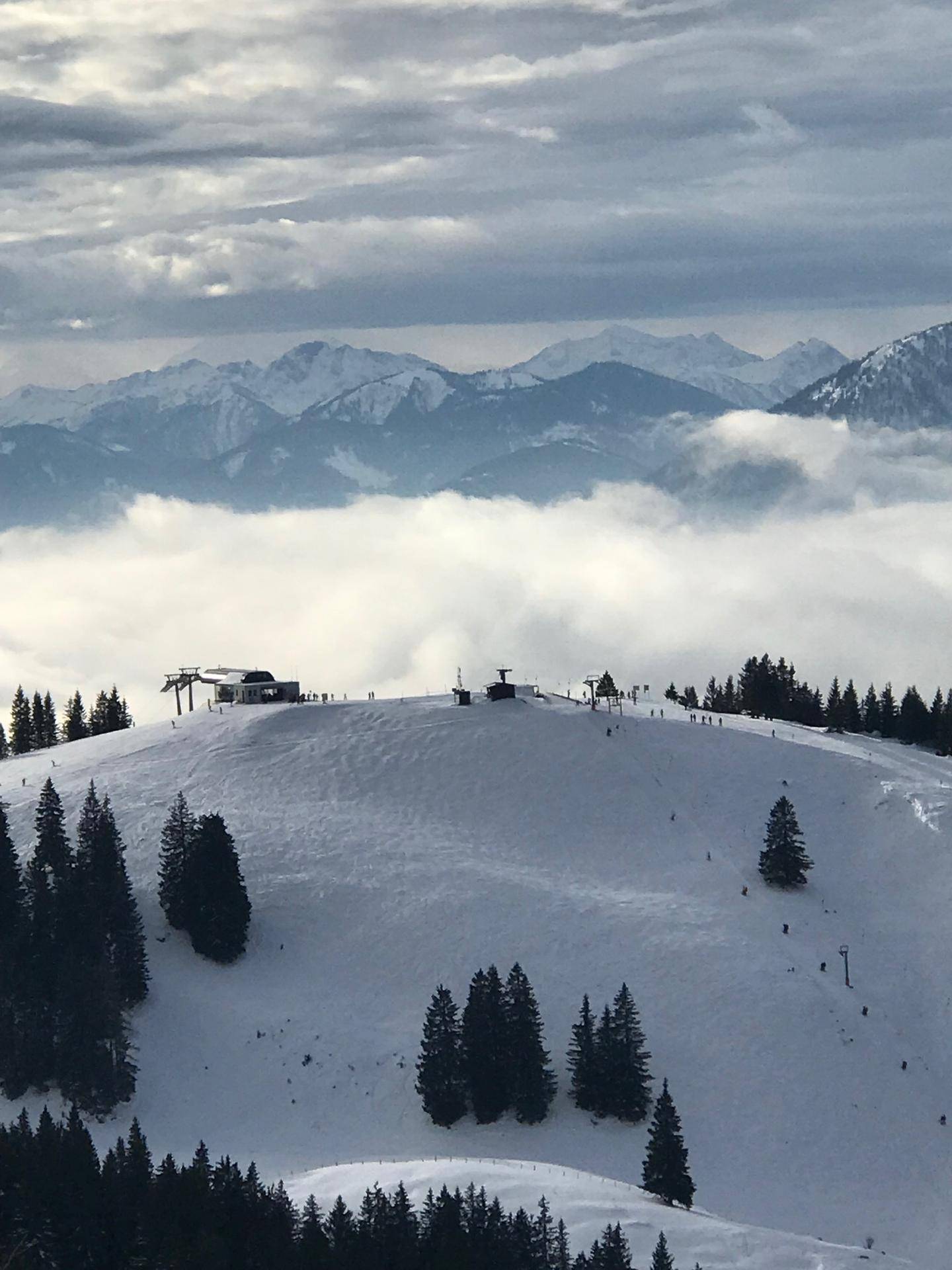 Panoramablick auf ein Skigebiet über den Wolken mit Berggipfeln im Hintergrund.
