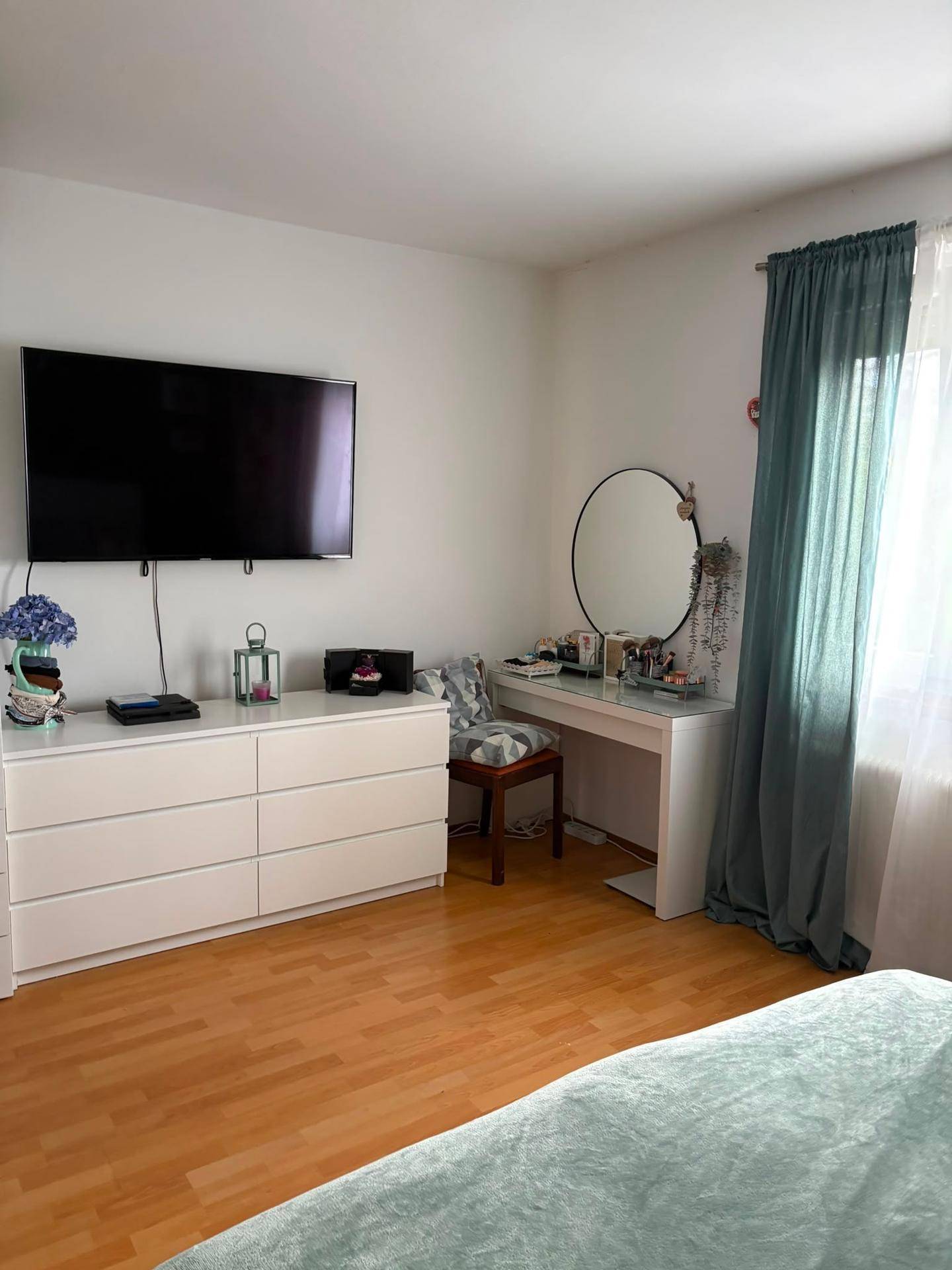 Schlafzimmer mit Holzboden, Kommode, Wandfernseher und Schminktisch am Fenster.