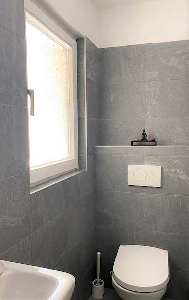 Separate Toilette mit Fenster, Wand-WC und grauen Fliesen.