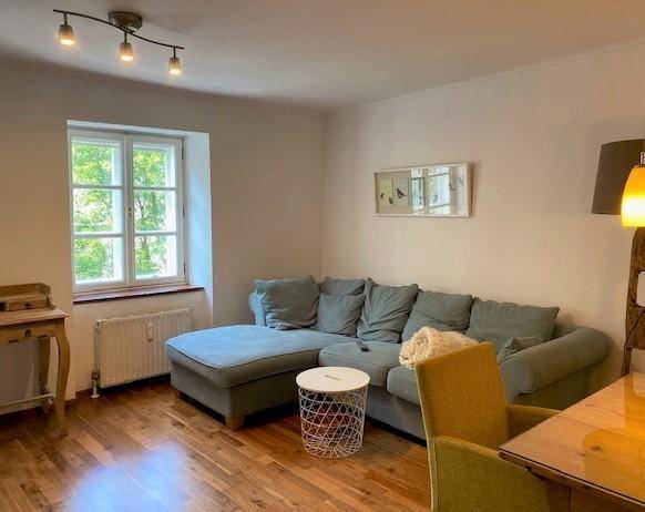 Gemütliches Wohnzimmer mit blauem Sofa, Holzboden und Fensterblick ins Grüne.