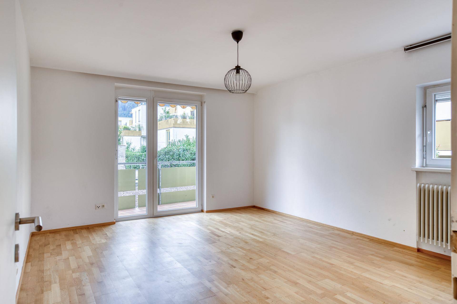 Geräumiges Zimmer mit hellem Parkettboden, großen Fenstern und Zugang zum Balkon.