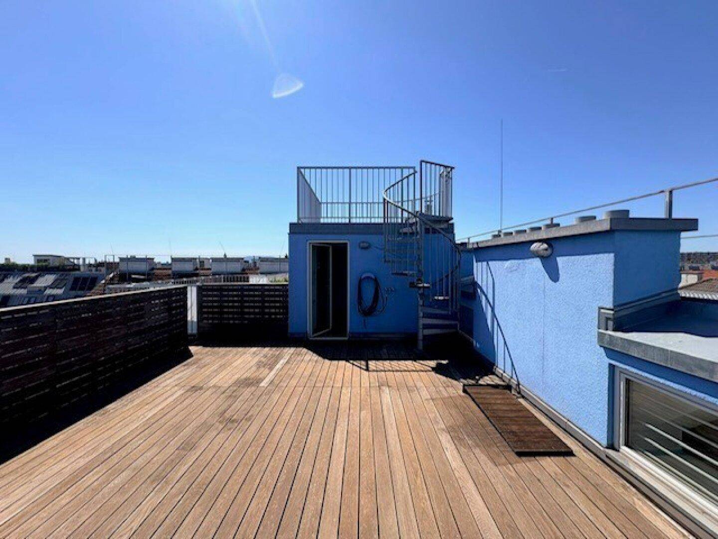 Weitläufige Dachterrasse mit Holzbelag, blauer Struktur und Wendeltreppe unter blauem Himmel.