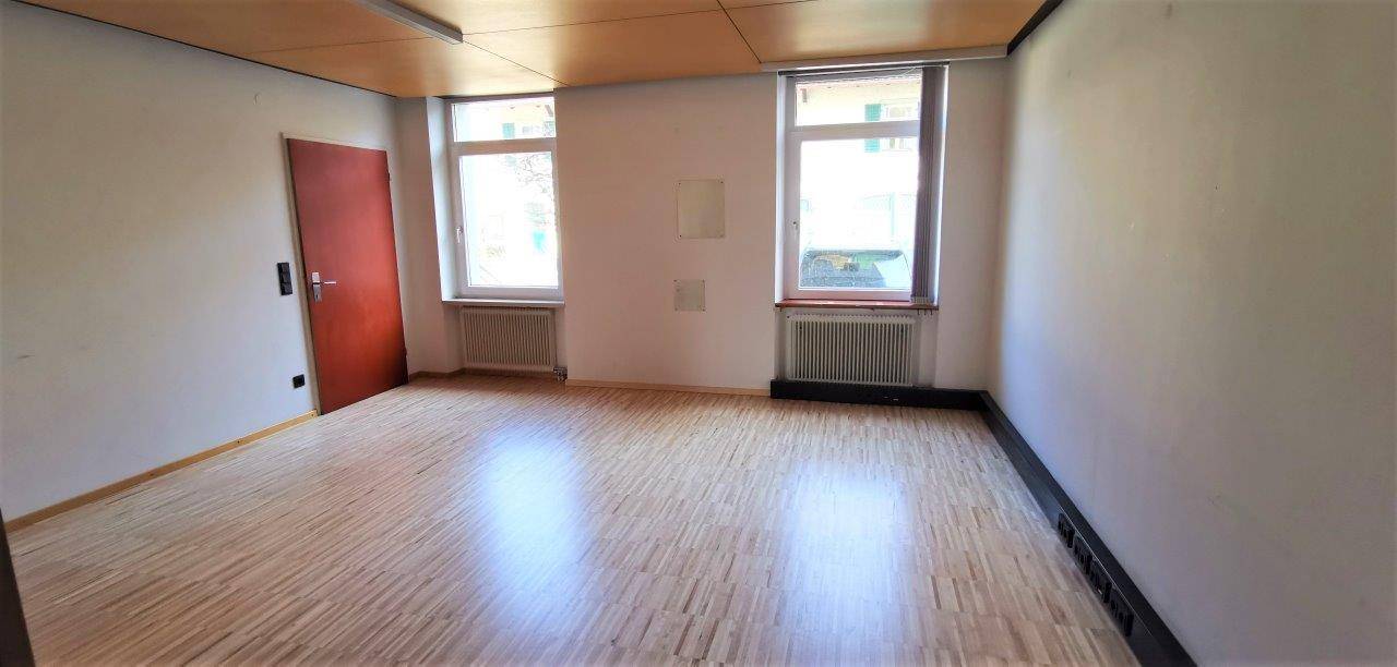 Heller Raum mit Holzdeckenverkleidung, Parkettboden und zwei Fenstern mit Heizkörpern darunter.