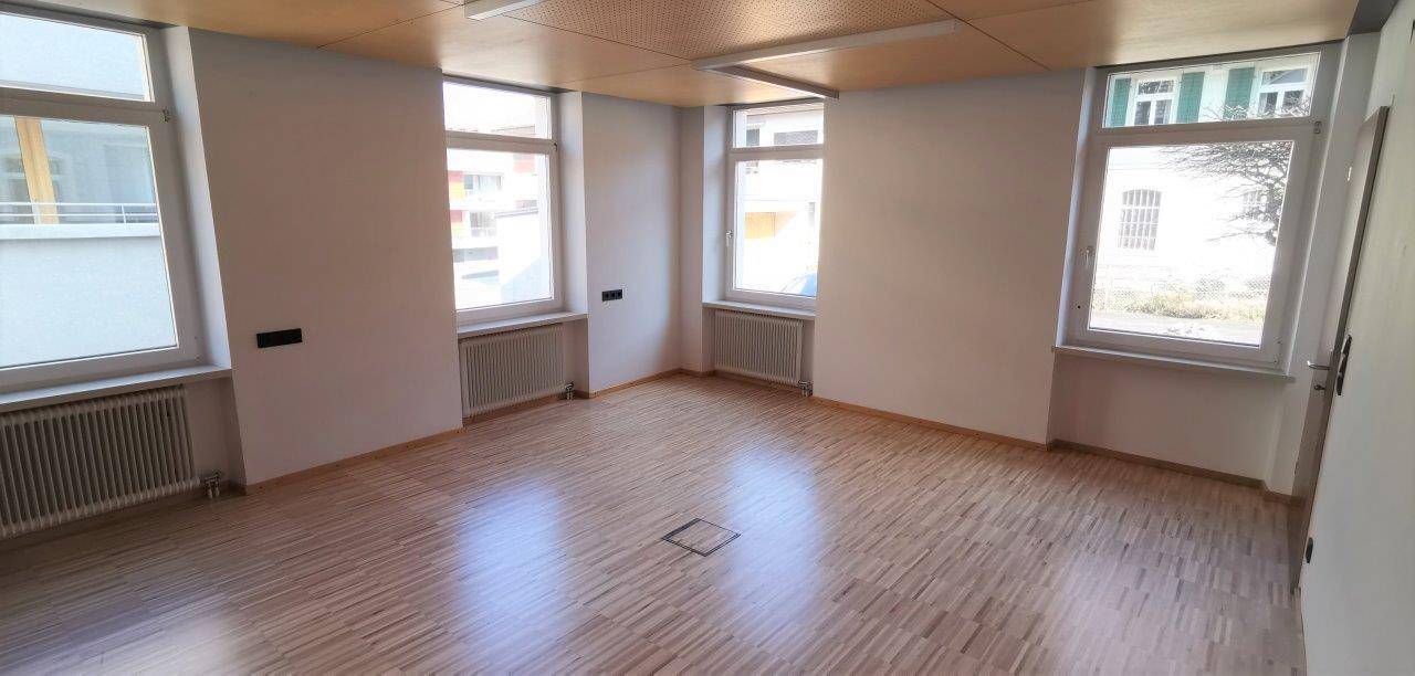 Großer, heller Raum mit mehreren Fenstern, Holzdeckenverkleidung und Parkettboden.