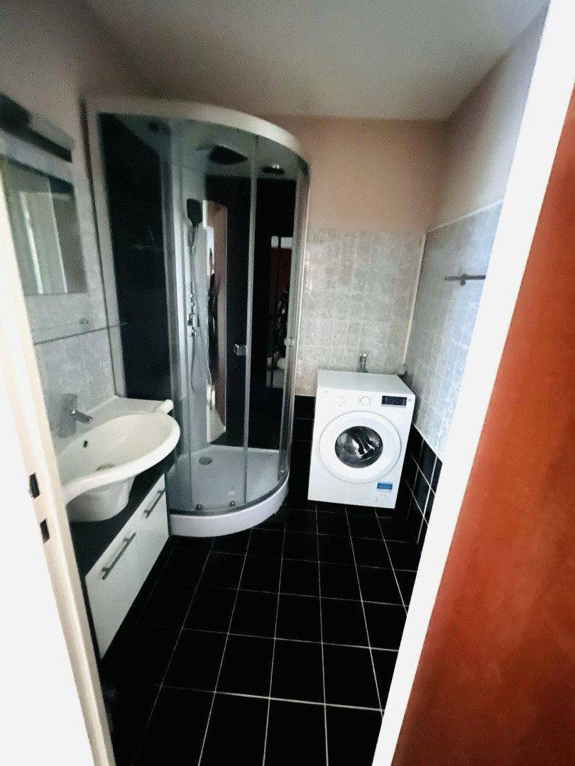 Badezimmer mit Eckduschkabine, Einzelwaschbecken und Waschmaschine auf schwarzen Fliesen.