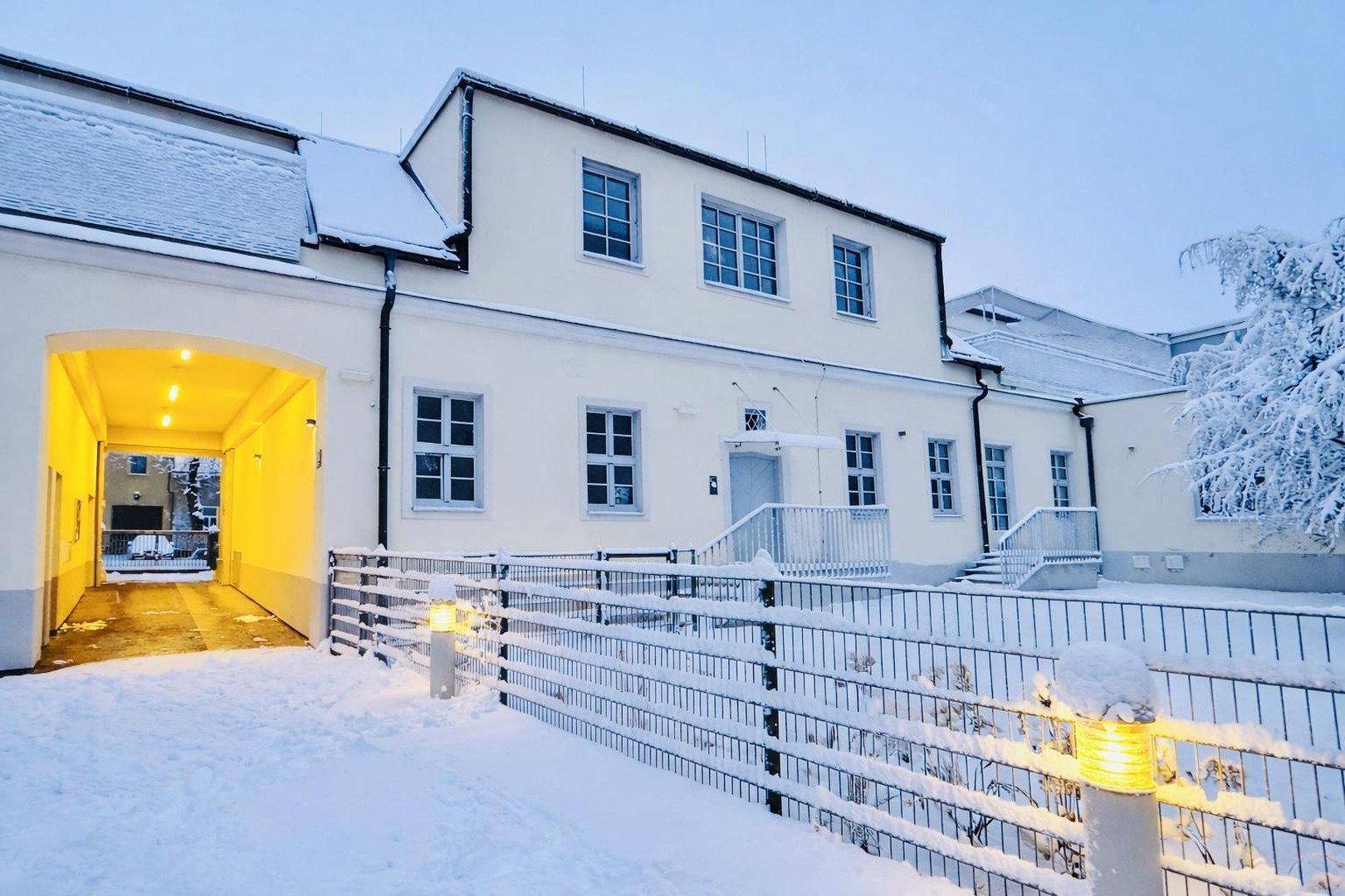 Rückansicht des Gebäudes im Winter mit verschneitem Hof und beleuchtetem Durchgang.