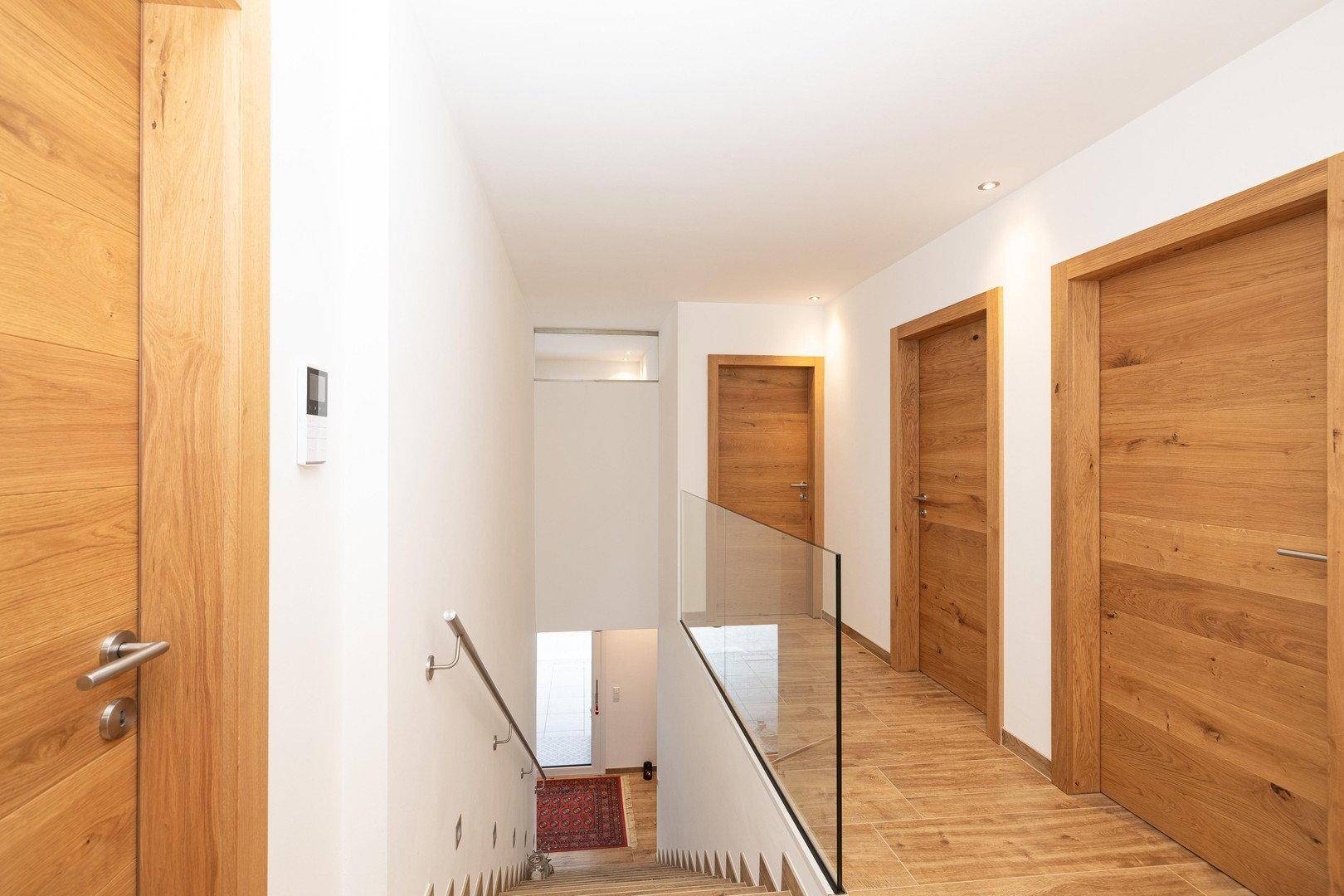 Heller Flur mit Holztreppe, Glasgeländer und mehreren Holztüren zu den Zimmern.